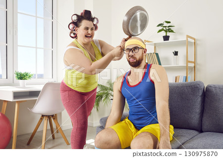 Funny, crazy housewife having fun and hitting her husband on head with frying pan 130975870