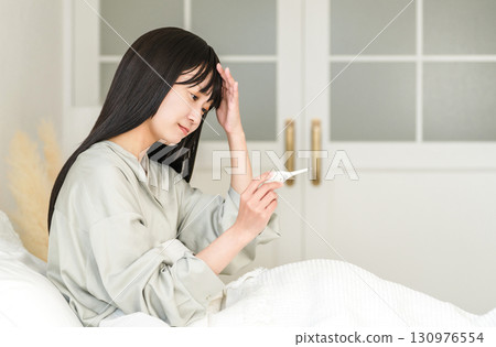 Unwell young Asian woman taking her temperature in bed (fever, cold) 130976554