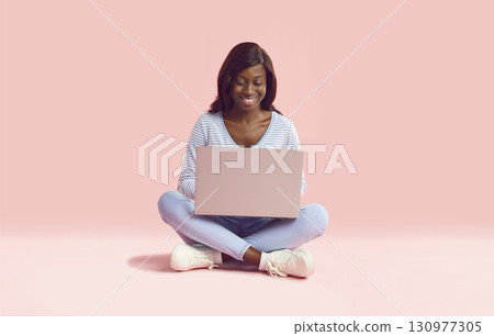 Happy young African American woman sitting on pink background and using laptop computer Happy young African American woman sitting on pink background and using laptop computer 130977305