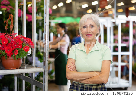 Successful mature woman, floral shop owner, standing near rack with petunias 130978259