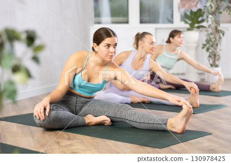 Yoga class of relaxed sports women performing Head to Knee Forward Bend Pose, or Janu Sirsasana after practicing together in gym 130978425