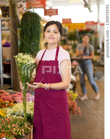 Middle-aged saleswoman holding bundle of small chamomiles in open-air plants market Middle-aged saleswoman holding bundle of small chamomiles in open-air plants market 130978593