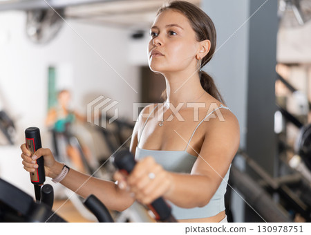 Slender athletic girl during cardio exercises on elliptical simulator in fitness center. 130978751