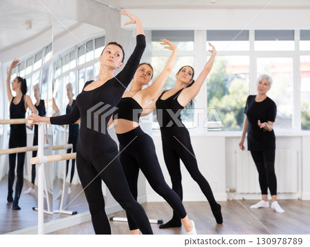 Mature teacher conducts class with girls in ballet studio, observes doing Batman exercise. 130978789