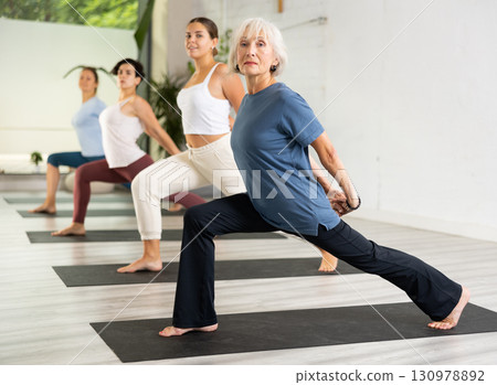 Active mature European woman practicing Virabhadrasana 1 - Warrior pose, indoor class with women, studio background 130978892