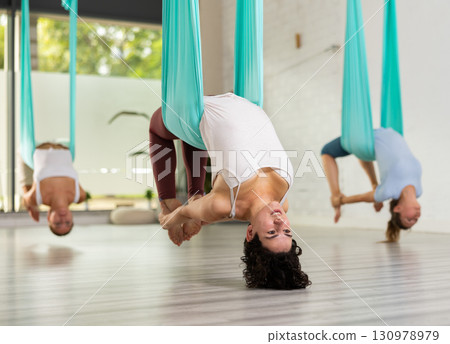 Full length side view portrait of woman hanging from silk hammock. Girl in elegant position practicing aerial yoga Full length side view portrait of woman hanging from silk hammock. Girl in elegant position practicing aerial yoga 130978979
