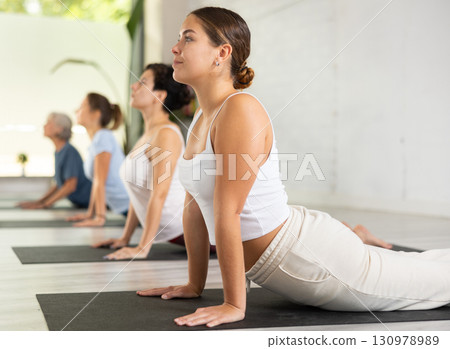 European young female practicing Bhujangasana or Cobra position during group workout at fitness studio 130978989