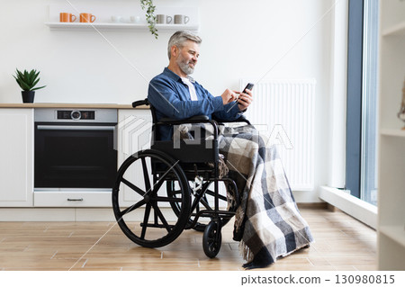Mature man in wheelchair with plaid blanket using smartphone indoors in bright modern kitchen 130980815