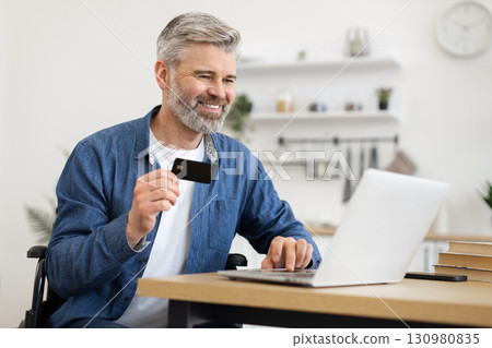 Middle-aged bearded man seated in wheelchair using laptop and bank card for online payment, demonstrating inclusivity, technology accessibility, online shopping convenience. 130980835