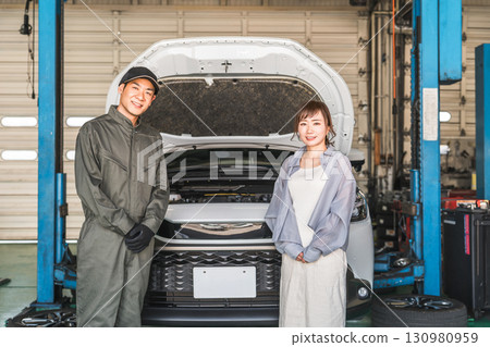 修理店裡,一名男性機械師和一名女顧客站在一輛被舉升的汽車前 修理店裡,一名男性機械師和一名女顧客站在一輛被舉升的汽車前 130980959