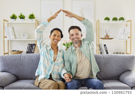 Happy couple celebrating relocation making hand roof above heads sitting on sofa in living room. 130981793