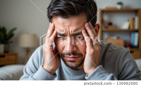 Man with headache holding head in pain at home, stressed and frustrated expression, indoor lifestyle, closeup portrait 130982000