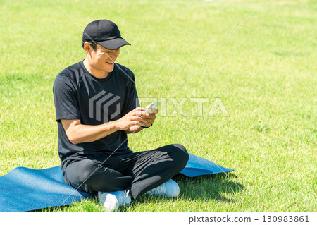 Asian man in sportswear sitting on the grass in the park and using his smartphone Asian man in sportswear sitting on the grass in the park and using his smartphone 130983861