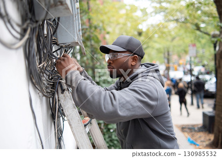 African American electrician in safety glasses laying wires outside a building on a city street 130985532
