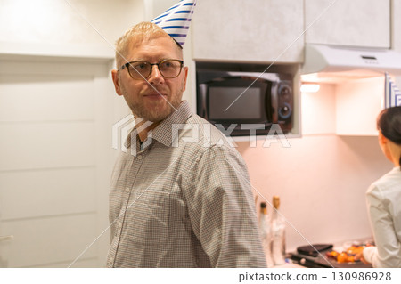 Here's a title attempting to follow all instructions: Birthday Man Kitchen Surprise Party Celebration: A man wearing a party hat is surprised in a kitchen during a birthday celebration. 130986928