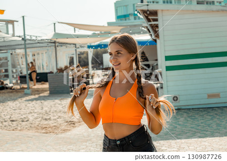 Woman Beach Summer Fashion: Smiling girl braids hair near beach kiosks, sunny day. 130987726