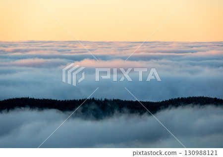 Winter inversion sunset with snow covered hills and forests rising above a sea of clouds, captured from above, with warm evening light painting the sky in glowing tones. 130988121