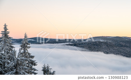 Winter inversion sunset with snow covered hills and forests rising above a sea of clouds, captured from above, with warm evening light painting the sky in glowing tones. 130988123