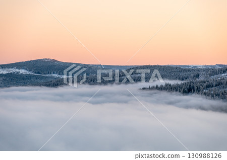Winter inversion sunset with snow covered hills and forests rising above a sea of clouds, captured from above, with warm evening light painting the sky in glowing tones. 130988126