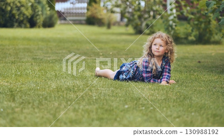 Blonde girl resting on verdant lawn, relaxing amid blooming garden surroundings with cheerful family atmosphere 130988192