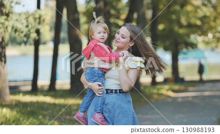 Cheerful mom raising laughing child high during playful outdoor moment amid green park landscape bathed in warm sunlight Cheerful mom raising laughing child high during playful outdoor moment amid green park landscape bathed in warm sunlight 130988193