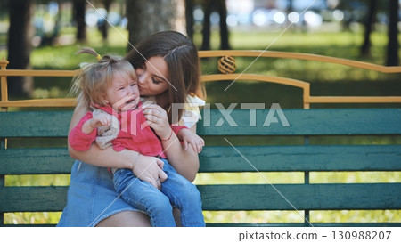 Young mother calming down her crying toddler daughter while sitting on a bench in a park during a sunny day 130988207
