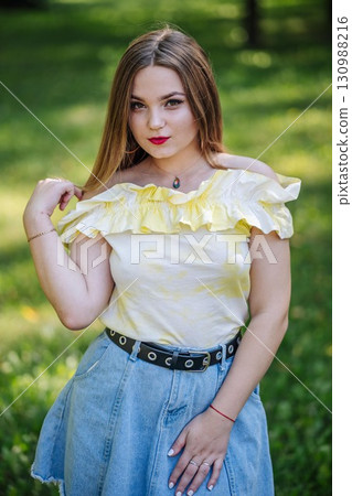 Young woman standing confidently in sunlit park, wearing stylish summer outfit amid verdant landscape and bright daylight 130988216