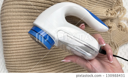 A woman holds a lint remover against a warm sweater with bobbles, removing pilling and caring for clothes. Preparing wardrobe for winter and autumn seasons, clothing care and maintenance A woman holds a lint remover against a warm sweater with bobbles, removing pilling and caring for clothes. Preparing wardrobe for winter and autumn seasons, clothing care and maintenance 130988431