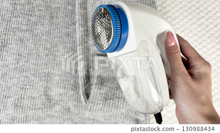A woman holds a lint remover against a warm sweater with bobbles, removing pilling and caring for clothes. Preparing wardrobe for winter and autumn seasons, clothing care and maintenance 130988434