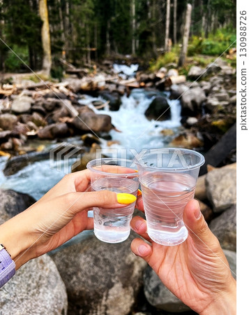 Two hands holding glasses of clear water, with a mountain river in the background. Two hands holding glasses of clear water, with a mountain river in the background. 130988726