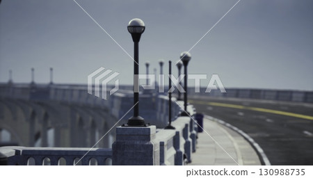 A series of lamp posts line the edge of a winding bridge, creating a peaceful atmosphere. The road appears empty, inviting a tranquil drive over the water below under a clear sky. 130988735