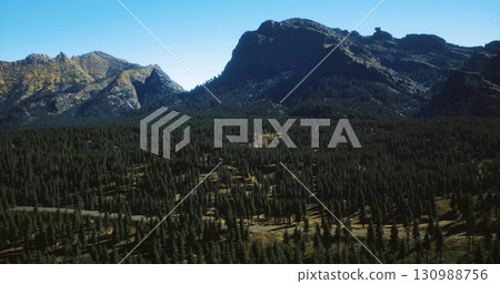 Expansive view of towering mountains surrounded by dense pine forests. Clear blue sky enhances the serene atmosphere of the natural landscape during daylight. 130988756