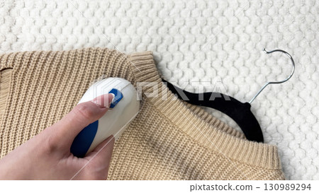A woman uses a fabric shaver to remove pilling from her warm autumn winter sweater. Clothes care, wardrobe preparation for cold season, lint remover cleaning knitwear and cozy wool garments A woman uses a fabric shaver to remove pilling from her warm autumn winter sweater. Clothes care, wardrobe preparation for cold season, lint remover cleaning knitwear and cozy wool garments 130989294
