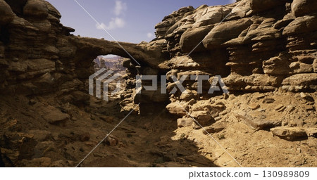A stunning view of a natural rock arch amidst rugged terrain in a sunlit desert. The dry landscape features layered rock formations and unique shapes, creating an otherworldly atmosphere. 130989809