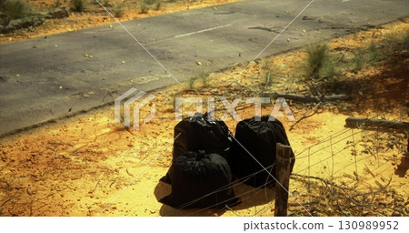 In a remote desert setting, three bags filled with trash sit abandoned next to an empty road. Sunlight casts long shadows and highlights the contrasting colors of the dry terrain. 130989952