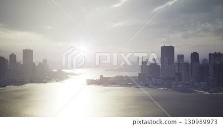 A stunning view of a city skyline reflects in the calm waters as the setting sun casts a silvery light. Skyscrapers loom majestically against a backdrop of cloudy twilight. 130989973