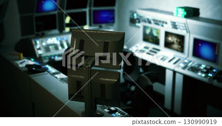 A dimly lit control room features multiple monitors displaying various data and graphics. The sophisticated setup suggests a hub for managing complex operations or systems. 130990919