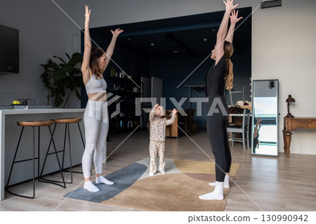 Two women in sportswear and a child practicing yoga together at home, standing with arms raised upward, healthy family training, stretching, exercising, indoor fitness and wellness routine 130990942