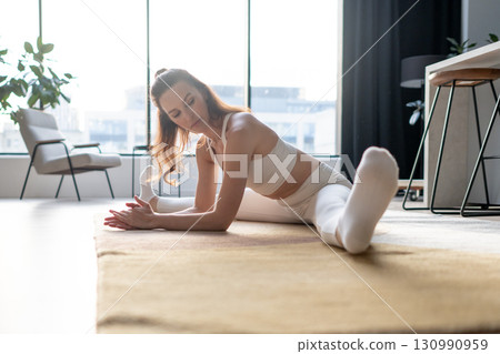 A woman in white yoga clothes stretches forward indoors, resting her forearms toward one extended leg, practicing hamstring opener, posture training, and flexibility improvement in daily exercising 130990959