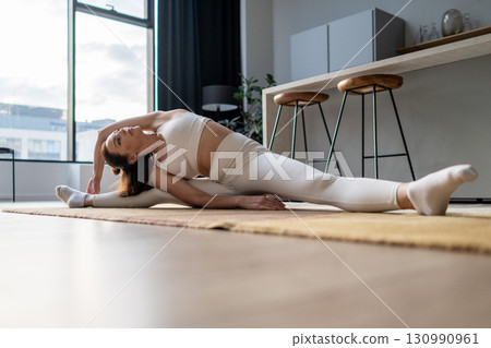 A woman in white sportswear performs seated side stretch on the floor, bending over one extended leg with arm overhead, practicing flexibility, body balance, and mindful breathing during a home yoga 130990961