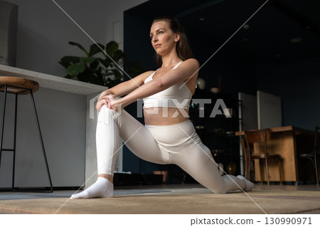 A woman in white activewear practices a yoga side lunge indoors, bending one leg and keeping the other extended, focusing on hip flexibility, joint mobility, and mindful breathing during home session 130990971