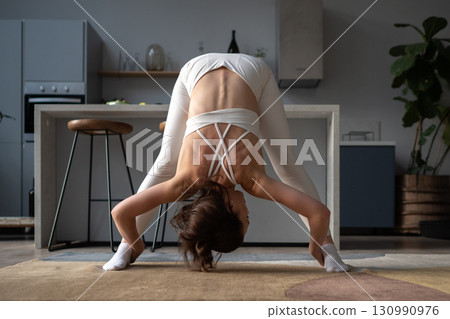 A woman in white activewear practices a yoga wide-legged forward bend indoors, leaning down with hands holding her feet, improving hamstring flexibility, spine elongation, and balance home workout 130990976