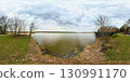 Panoramic 360-degree view of a lakeshore surrounded by early spring trees with sparse leaves. A playground is visible on the right, and a sandy beach with boats on the left. The sky is filled with dra 130991170