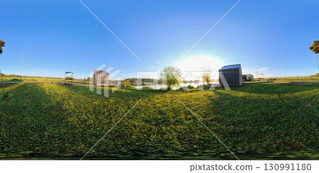 360-degree panoramic HDRI image of a serene countryside scene at sunrise. A wooden boardwalk stretches between grassy fields and small lakeside cabins. 130991180