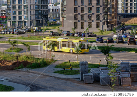 A vivid yellow bus travels along a circular route in a bustling neighborhood. Nearby, modern buildings stand tall amidst parked cars and greenery A vivid yellow bus travels along a circular route in a bustling neighborhood. Nearby, modern buildings stand tall amidst parked cars and greenery 130991799