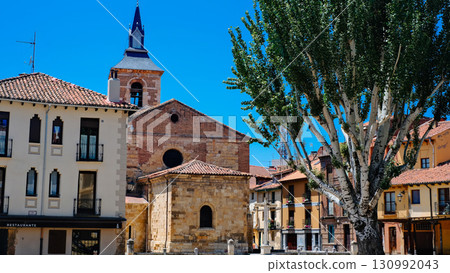 A picturesque, sun-drenched town square in Leon, Spain, featuring a historic stone church with a dark slate bell tower 130992043