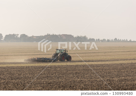 Tractor pulls plowing equipment across wide harvested farmland under pale hazy sky. Dust rises as soil is prepared for next agricultural season in rural countryside 130993480