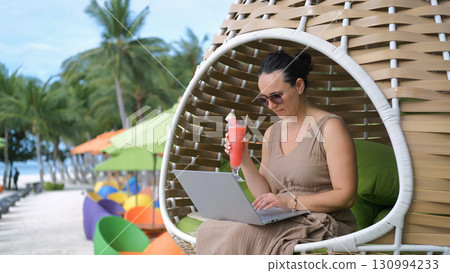 Young woman relaxes in a woven chair working on her laptop sipping a watermelon shake and enjoying the beach scene  130994233
