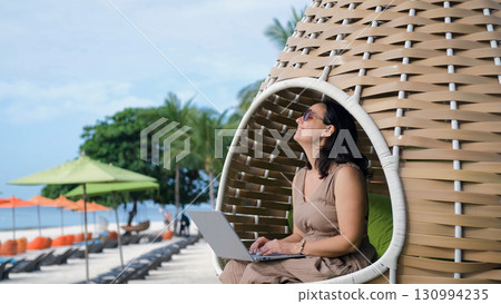 A young woman relaxes in a woven chair working on her laptop while enjoying the tropical beach embracing freelance lifestyle A young woman relaxes in a woven chair working on her laptop while enjoying the tropical beach embracing freelance lifestyle 130994235