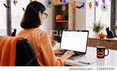 woman wearing glasses uses a laptop while sitting at a table in a room decorated for halloween 130994500